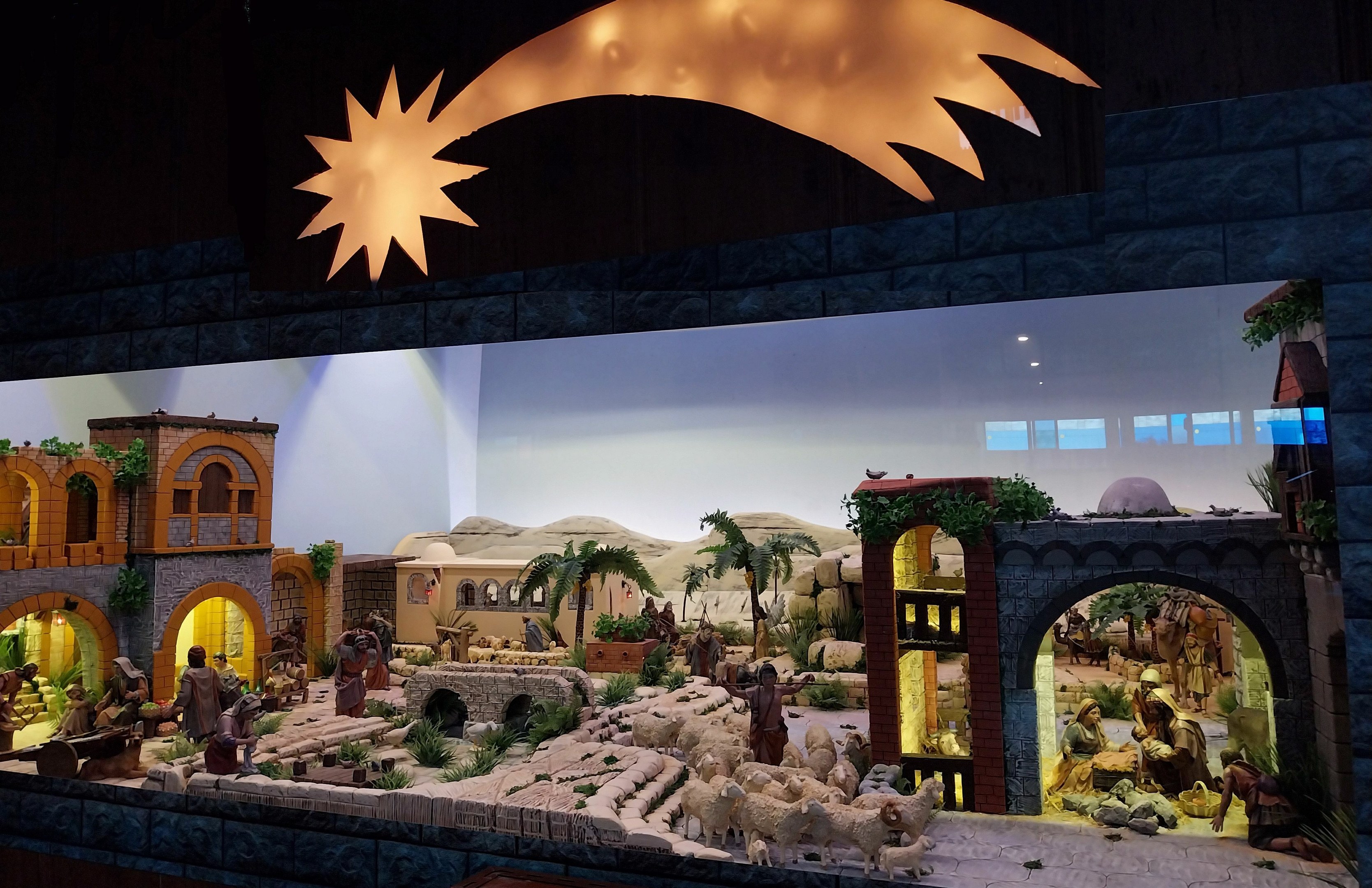 Eine Krippe-Ausstellung im Museum mit Menschen, Tieren, Häusern, Bäumen und anderen Gegenständen sowie einer beleuchteten Hintergrundgestaltung.