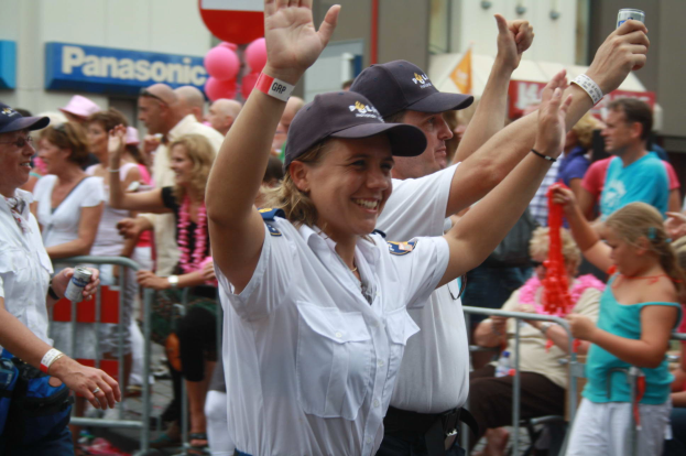 Eine Gruppe von Menschen, einige mit Mützen und Dosen haltend, steht und sitzt vor einer Menge, mit Geländern, Luftballons und einem Gebäude mit Namensschildern im Hintergrund.