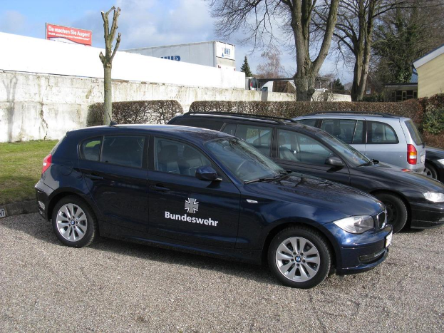 Mehrere Autos sind auf einem Weg geparkt, der von grünem Gras, Büschen und Bäumen umgeben ist. Im Hintergrund sind Gebäude, ein Schild mit Text und ein Logo zu sehen.