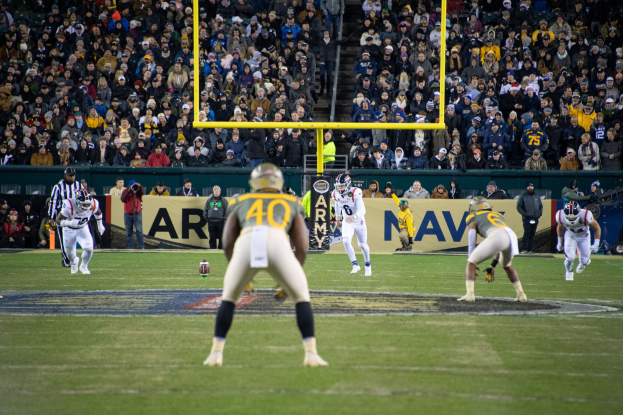 Fußballspieler in Sportkleidung und Helmen konkurrieren auf einem Feld mit Zuschauern in den Rängen, Pfosten und sichtbarer Beschilderung im Hintergrund.