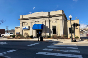 Ein Gebäude mit einem blauen Vordach an einer Straßenecke, umgeben von Pflanzen, Laternen, Fahrzeugen, Bäumen und Strommasten mit Drähten, unter einem bewölkten Himmel, identifiziert als Champion Bank & Trust Co.