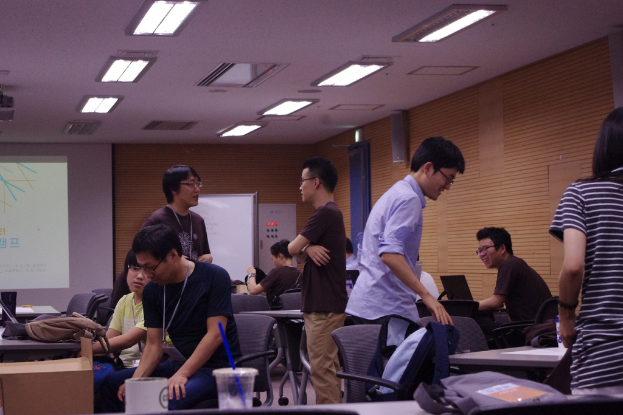 Ein Klassenzimmer mit Tischen, Stühlen, Laptops und Taschen; Menschen, die stehen und sprechen, ein Projektionsschirm, eine Holzwand und eine Tür im Hintergrund.