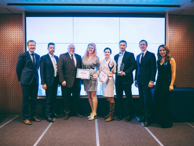 Gruppe von strahlenden Menschen mit Urkunden, Gewinner der Internationalen Business Awards 2018, vor einem Bildschirm mit einem Tisch und einem Wandobjekt im Hintergrund.