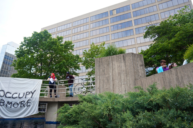 Menschen auf einer Brücke mit einem Banner und einem Fahrrad, Wasser darunter, Bäume, Gebäude und Himmel im Hintergrund.