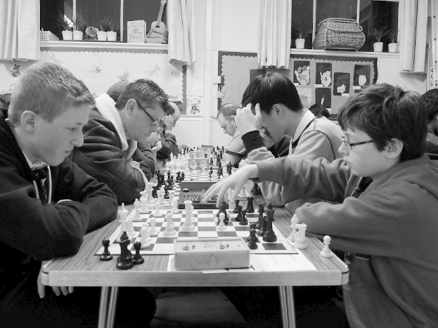 Eine Gruppe von Menschen sitzt an Tischen und spielt Schach, mit Schachbrettern, Münzen und Timern auf den Tischen. Ein Junge in der rechten Ecke trägt eine Brille, und im Hintergrund ist eine Wand mit Vorhängen, einer Gitarre, Körben, Gläsern und Gemälden zu sehen.