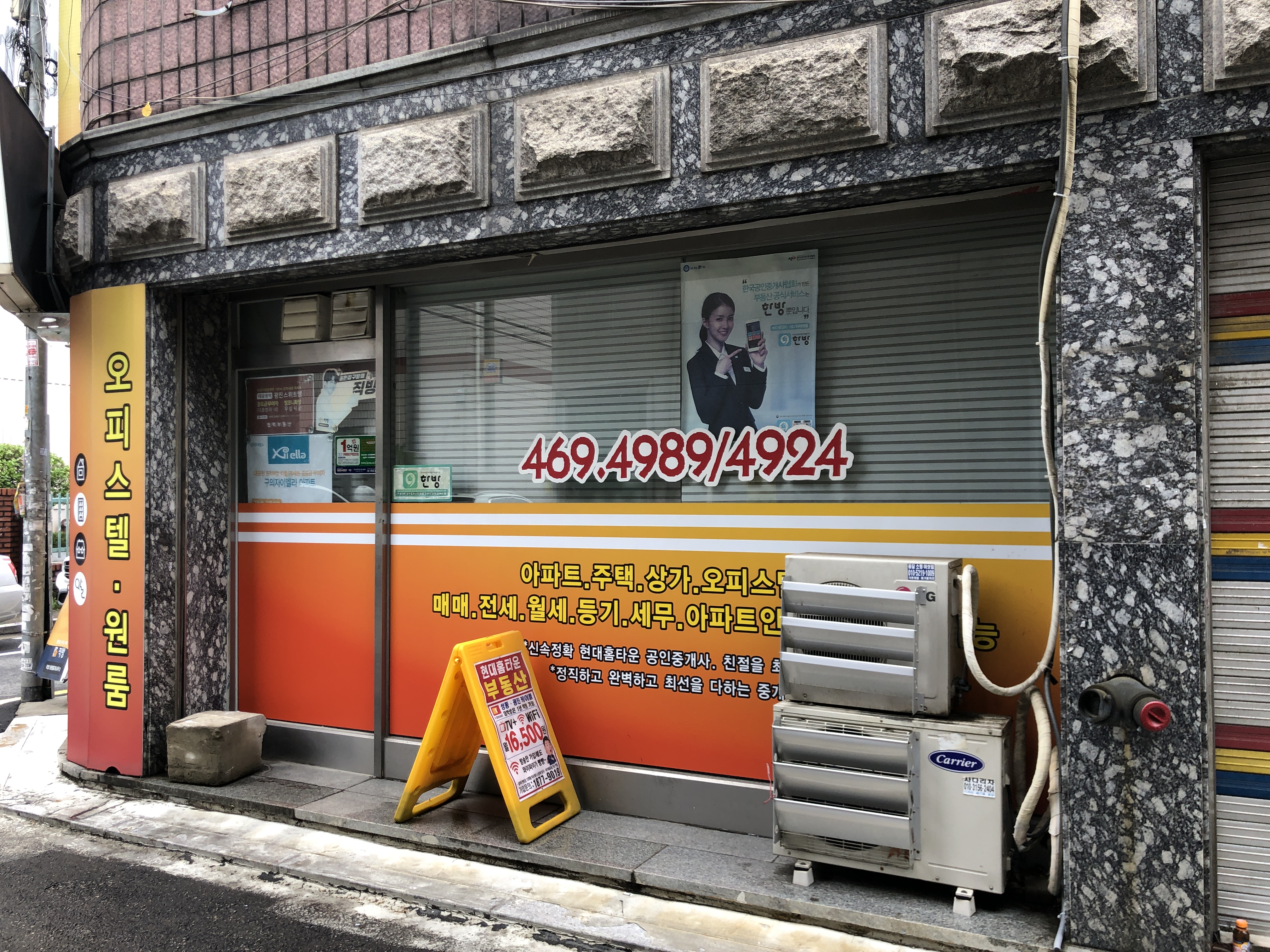 Ein Gebäude mit einer Tafel, die "Koreanische Klimatechnik-Service" lautet, einem Rollladen, einer Tafel mit Text und einem Personenbild, verschiedenen Geräten auf dem Boden, Pfählen, Bäumen, einem Auto auf der Straße und Drähten unter einem bewölkten Himmel.