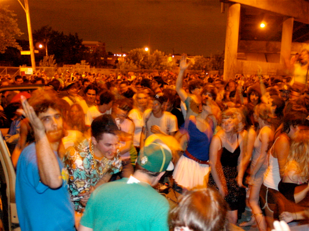 Menschen tanzen vorne, während im Hintergrund ein Konzert mit Lichtern, Pfählen, Bäumen und anderen Objekten stattfindet.