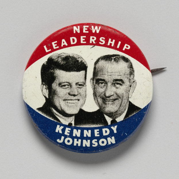 Eine weiße Oberfläche mit einem Ansteckbutton mit zwei Bildern von Kennedy und Johnson, darunter die Wörter "Neue Führung".