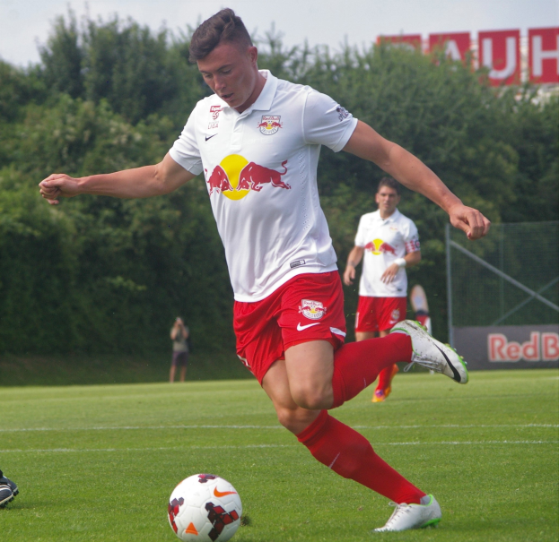 Ein Mann in einem Fußballtrikot kickt einen Ball auf einem grünen Rasen mit Bäumen und einem klaren blauen Himmel im Hintergrund, mit einem "RB Leipzig"-Schild im Bild.