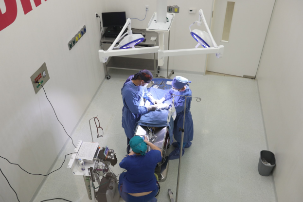 Eine Gruppe von Chirurgen führt einen Eingriff in einem Operationssaal durch, wobei ein Patient auf einem Operationstisch umgeben von medizinischer Ausrüstung liegt, eine textbedeckte Wand auf der linken Seite, ein Objekt auf dem Boden auf der rechten Seite und verschiedene medizinische Geräte im Hintergrund.