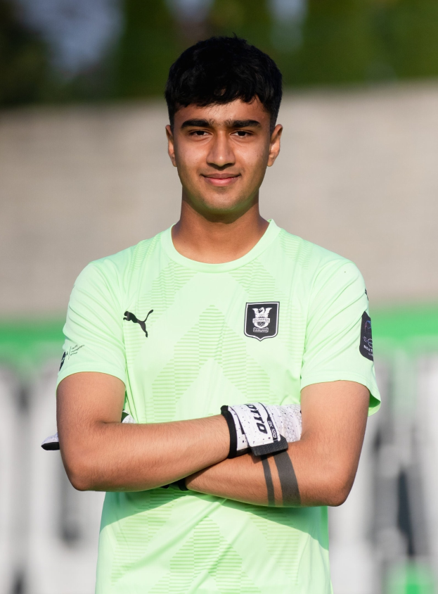 Ein junger Mann in grünem T-Shirt steht auf einem Fussballfeld und schaut mit verschräckten Armen und einem Lächeln in die Kamera, mit einem unscharfen Hintergrund.