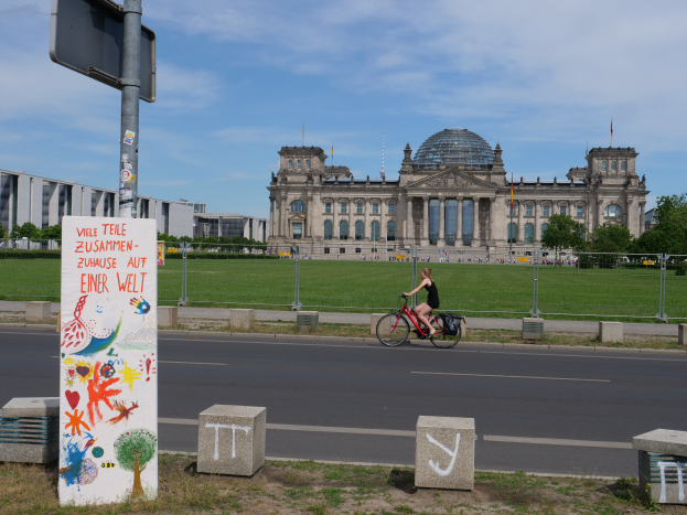 Eine Person in einem schwarzen Kleid fährt mit dem Fahrrad eine Straße vor dem Reichstaggebäude in Berlin, Deutschland, entlang, mit Gebäuden, Bäumen und Menschen im Hintergrund unter einem blauen und weißen Himmel.