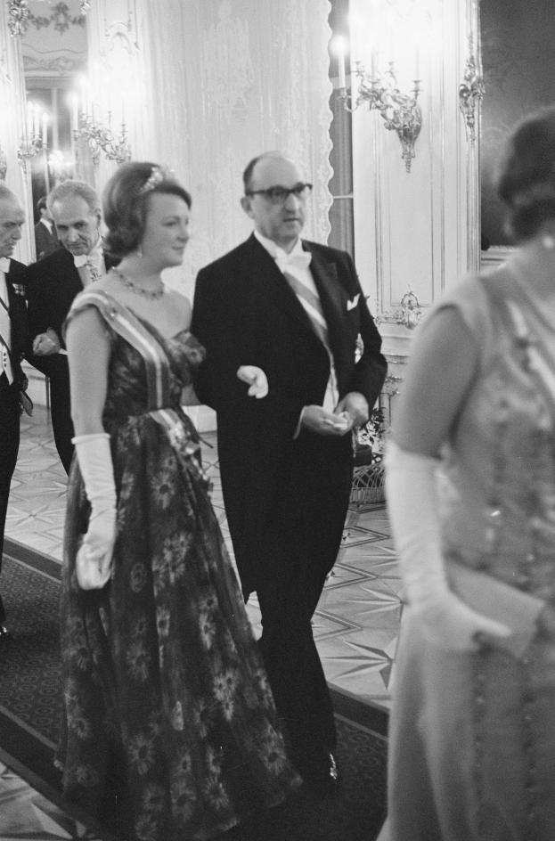 Ein Mann in einem schwarzen Blazer, weißem Hemd und schwarzen Hosen sowie eine Frau in einem eleganten Kleid gehen einen Flur entlang, umgeben von einer Gruppe von Menschen, die auf dem Boden stehen, während einige Wandlampen eine gemütliche Atmosphäre schaffen.