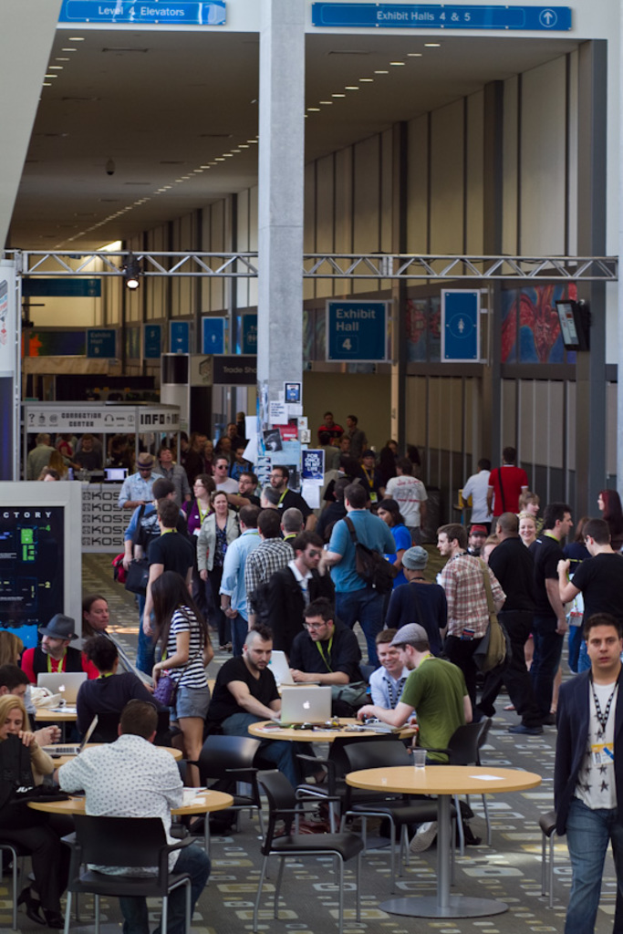 Ein belebter Flughafenvorhalle mit wartenden Menschen, einige stehen und andere arbeiten, Richtungsschilder an der rechten Wand und darüber sowie von der Decke hängende Lampen.