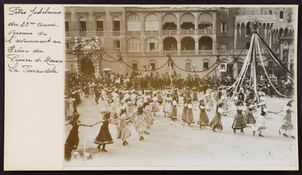 Ein Schwarz-Weiß-Foto einer Gruppe von Menschen in traditioneller Kleidung, die vor einem Gebäude mit Fenstern, Bögen, Säulen und Geländern tanzen, während eines Karnevals in Paris, mit einem Pfahl mit daran gebundenen Gegenständen auf der rechten Seite und Text auf der linken Seite.