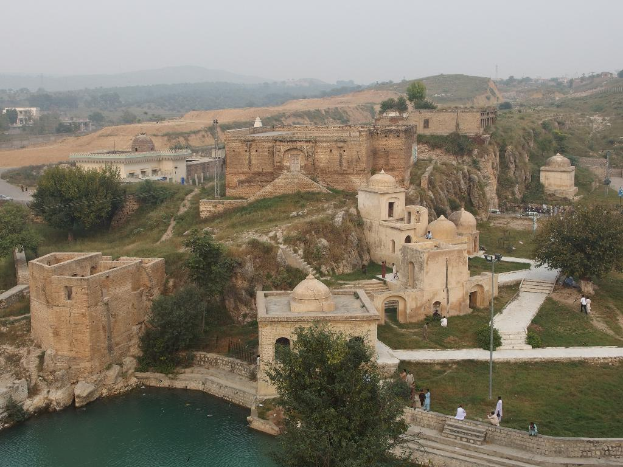 Eine Landschaft mit zahlreichen Burgen und Gebäuden, umgeben von Gras und Bäumen, mit einem Teich im Vordergrund, Hügel im Hintergrund und einem Himmel darüber.