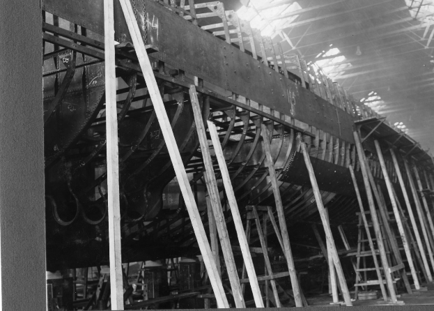 Schwarzes und weißes Foto eines Boots in der Fertigung in einer Fabrik, mit Holzpfählen, Leitern und anderen Gegenständen auf dem Boden und einer Decke darüber.