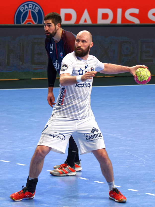 Zwei Männer beim Handballspielen auf einem Platz, einer hält den Ball, im Hintergrund eine Tafel mit "Paris Saint-Germain vs Paris Saint-Germain."
