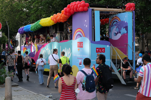 Eine Gruppe von Menschen, die eine Straße entlanggehen, neben einem Lastwagen mit bunten Luftballons, mit Schildern an Pfosten entlang der Straße und Bäumen und Gebäuden im Hintergrund, was auf eine Pride-Parade in Paris hindeutet.
