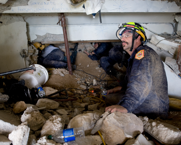 Ein Feuerwehrmann in einem Helm sitzt umgeben von Trümmern, einschließlich Steinen und Flaschen, mit einer teilweise sichtbaren Wand im Hintergrund, die wahrscheinlich den Namen des Gebäudes anzeigt, das von einem israelischen Luftangriff getroffen wurde.