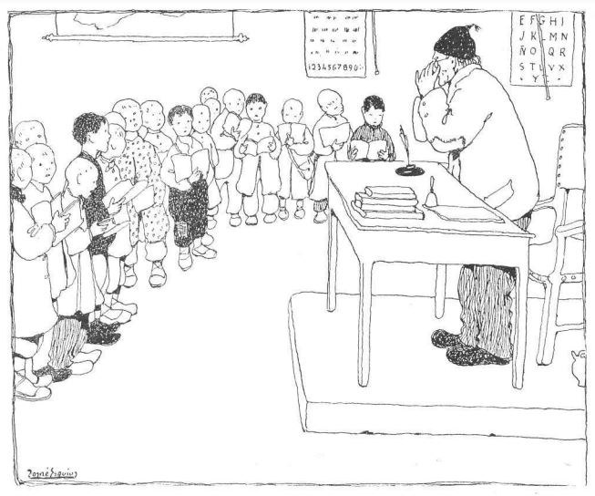 Ein Mann, mutmaßlich ein Lehrer, sitzt an einem Schreibtisch mit Büchern und Gegenständen vor einer Gruppe von Kindern, mit Papieren an der Wand hinter ihm.