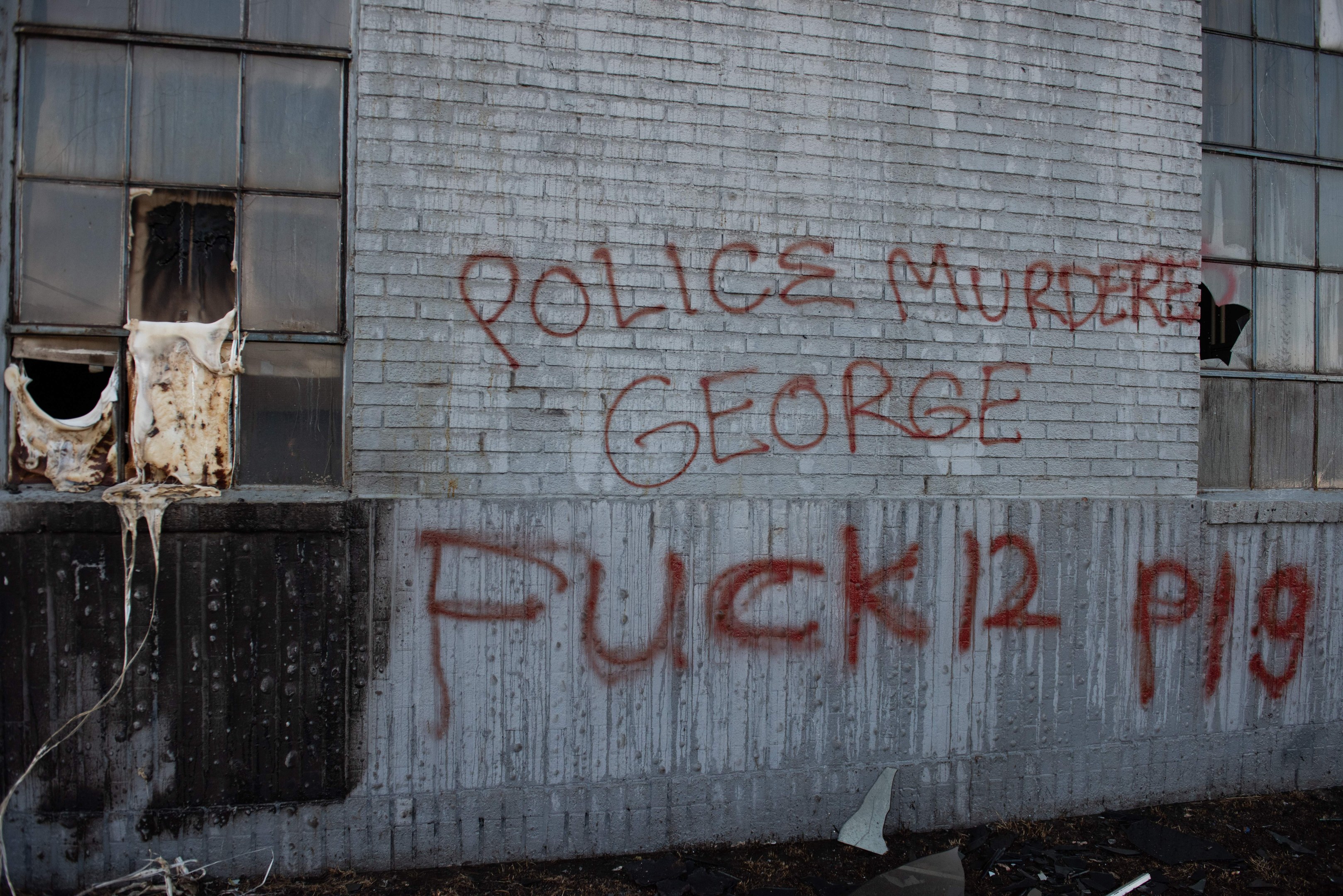 Graffiti an einer Gebäudewand mit der Aufschrift "Polizei ermordet George Fuck 12 P19" und Fenstern sowie verstreuten Gegenständen davor.