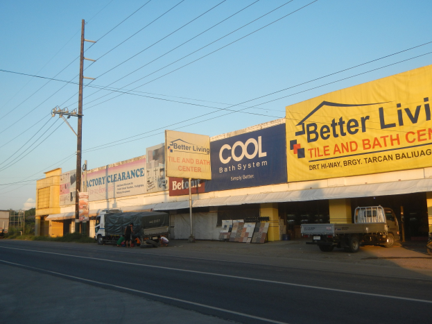 Eine belebte Stadtstraße mit Fahrzeugen, Fußgängern und einem Gebäude mit der Aufschrift "Better Living Tile and Bath Center" im Hintergrund unter einem bewölkten Himmel mit sichtbaren Strommasten und -drähten.