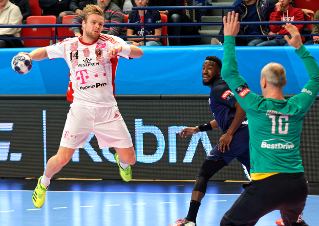 Eine Gruppe von Männern, die Handball auf einem Platz spielen, mit einem Ball in der Mitte der Aktion, während Zuschauer von Stühlen und Geländern in einem Stadion zusehen. Das Anzeigebrett im Hintergrund zeigt "Futsal-Weltmeisterschaft 2019 - Bayern München vs. Paris Saint-Germain".