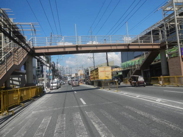 Eine Stadtstraße mit Fahrzeugen, eine Brücke mit Geländern und Treppen auf der linken Seite, Gebäude im Hintergrund, Strommasten mit Drähten und ein bewölkter Himmel.
