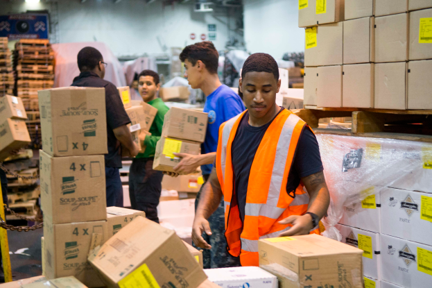 Eine Gruppe von Männern in orangefarbenen Westen und Sicherheitswesten lädt Kisten in ein Lagerhaus, mit mehreren Kartons auf der rechten Seite und einer Kette auf der linken Seite und einer Wand mit Lampen im Hintergrund.