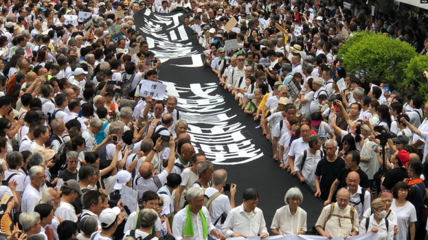 Eine große Gruppe von Menschen marschiert auf einer Straße, hält Protestschilder und Banner in der Hand, mit Grünpflanzen auf der rechten Seite und einem Gebäude im Hintergrund.