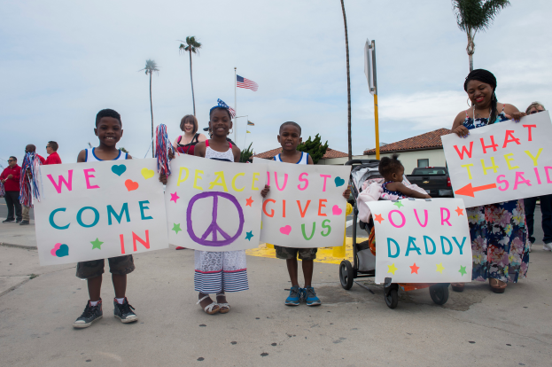 Eine Gruppe von Kindern hält ein Schild mit der Aufschrift "Wir kommen in Frieden, gebt uns unseren Daddy" und steht auf einer Straße umgeben von Bäumen, Pfählen, einer Fahne, einem Haus, Pflanzen und einem bewölkten Himmel, mit einer Frau, die einen Kinderwagen mit einem Baby vor einem Gebäude schiebt.