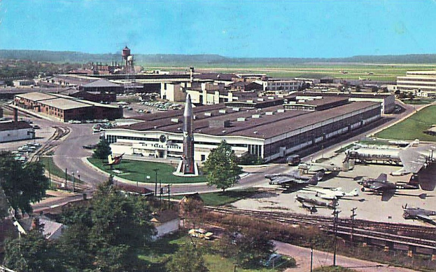 Altes Schwarz-Weiß-Foto eines Flughafens mit geparkten Flugzeugen, Gebäuden, Bäumen, Straßen, Fahrzeugen, Strommasten, Gras, Bahnschienen, Türmen, Hügeln und einem klaren Himmel, mit Text unten.