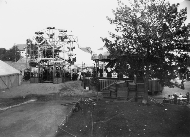 Ein Schwarz-Weiß-Foto eines Karnevals mit einem Riesenrad in der Mitte, umgeben von Menschen, Zelten, Bäumen und anderen Gegenständen, mit einer Straße unten.