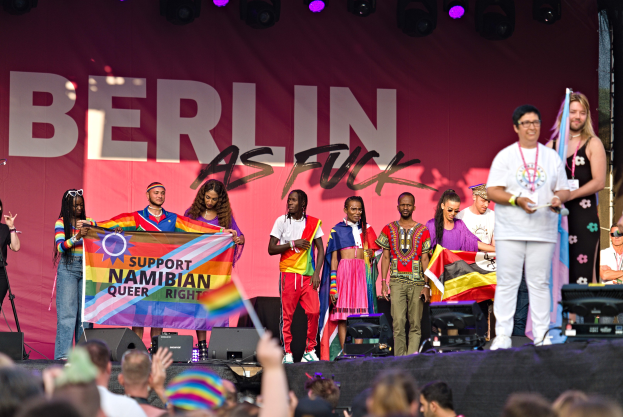 Gruppe von Menschen auf einer Bühne mit einem Banner, auf dem 'Berlin as Fuck' steht, mit Lautsprechern, Mikrofonen und einer Menge unten, die Hände hebt.