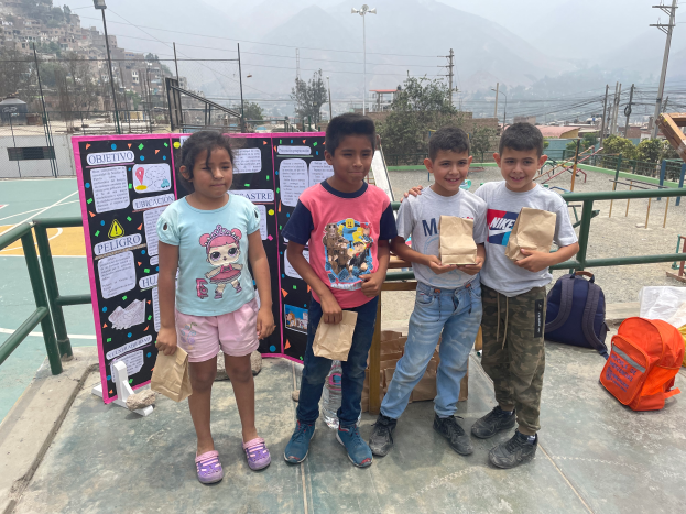 Eine Gruppe von Kindern steht auf einem Basketballfeld, jedes Kind hält eine Papiertüte in der Hand, mit Tüten und Gegenständen rechts, einer Texttafel links und einer Kulisse aus Zaun, Pfosten, Straßenlaternen, Bäumen, Gebäuden, Hügeln und Himmel.
