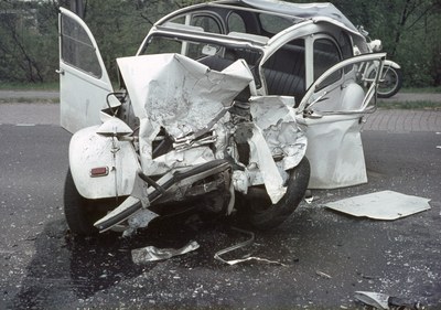 Ein schwerbeschädigt aussehender Pkw mit eingedrückter Frontpartie am Rödrand, umgeben von Bäumen und einem Gebäude im Hintergrund, mit einem weiteren Fahrzeug in der Ferne.
