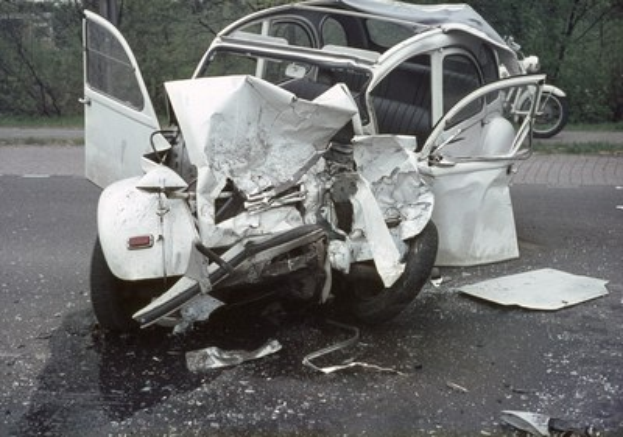 Ein schwerbeschädigt aussehender Pkw mit eingedrückter Frontpartie am Rödrand, umgeben von Bäumen und einem Gebäude im Hintergrund, mit einem weiteren Fahrzeug in der Ferne.