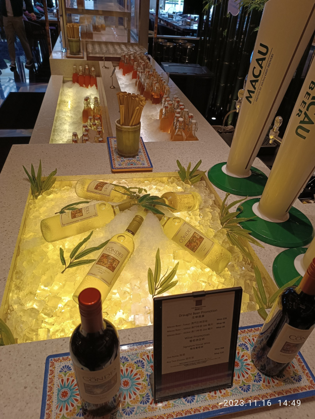 Ein Tisch mit Weinflaschen, Eiswürfeln, Pflanzen, Brettern und anderen Gegenständen vor dem Hintergrund von Menschen, Tischen, Stühlen und Glastüren beim Macau-Bierfestival.