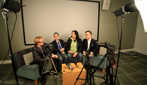 Eine Gruppe von drei Personen sitzt auf einem Sofa und gibt ein Interview, wobei eine Person in einem schwarzen Mantel und Hose links, eine Person in einem Mantel und blauer Krawatte mit einem Mikrofon in der Mitte und eine Person mit einem Lächeln rechts zu sehen ist. Ein Mikrofonrekorder befindet sich auf dem Tisch in der Mitte, Kameras sind auf der rechten und linken Seite und große Scheinwerfer sind vorhanden, sowie eine Projektionswand im Hintergrund.