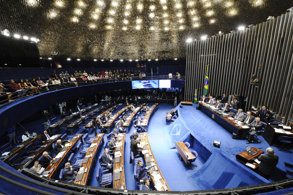 Eine Konferenzbühne mit sitzenden Personen an einem Tisch mit Papieren und Gegenständen, flankiert von Fahnen, einer Skulptur im Hintergrund und einem Publikum, das an Tischen sitzt und hinter einem Geländer mit Bildschirmen und Deckenleuchten.