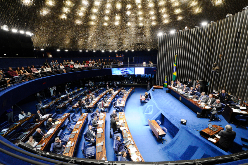 Eine Konferenzbühne mit sitzenden Personen an einem Tisch mit Papieren und Gegenständen, flankiert von Fahnen, einer Skulptur im Hintergrund und einem Publikum, das an Tischen sitzt und hinter einem Geländer mit Bildschirmen und Deckenleuchten.