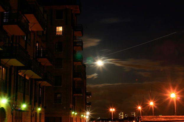 Eine nächtliche Stadtansicht mit einem prominenten Gebäude im Vordergrund, das zahlreiche Fenster und Balkone aufweist, sowie mehreren Straßenlaternen auf der rechten Seite.