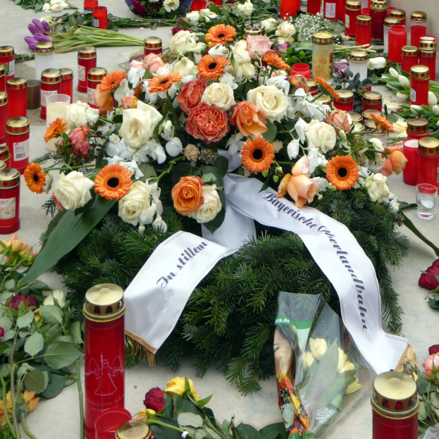 Mahnmal in Berlin mit Blumen, Kerzen und Blättern vor einer Wand.