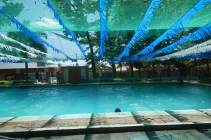Ein großes Schwimmbad mit blauen und grünen Bändern, die von der Decke hängen, umgeben von Häusern, Bäumen und einem klaren blauen Himmel, mit Menschen, die im Wasser und in der Sonne genießen.