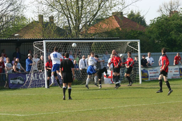 Fußballspieler spielen auf einem Feld mit einem Tor im Hintergrund, während Zuschauer zuschauen; Bäume und Häuser sind im Hintergrund zu sehen.