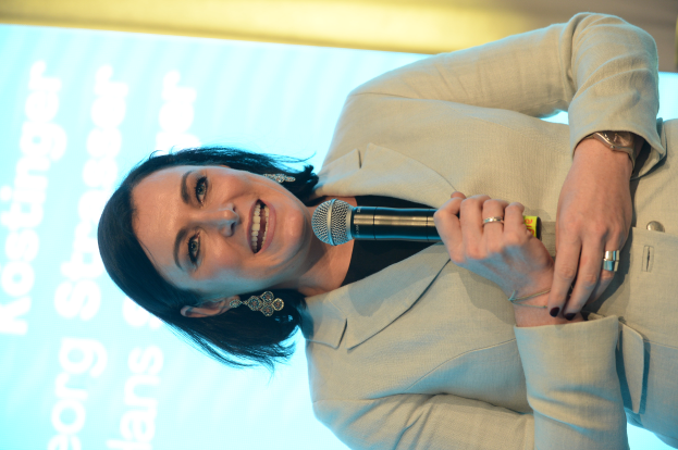 Eine Frau in einem beigen Anzug, mit einem Dutt und einem strahlenden Lächeln, hält ein Mikrofon und hat einen Bildschirm im Hintergrund.