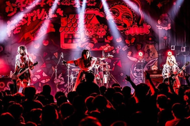 Ein Live-Auftritt der japanischen Rockband Korn auf der Bühne mit Musikern, die Gitarren spielen und in Mikrofone singen, ein beleuchtetes Wandgemälde im Hintergrund und eine sichtbare Menge im Publikum.