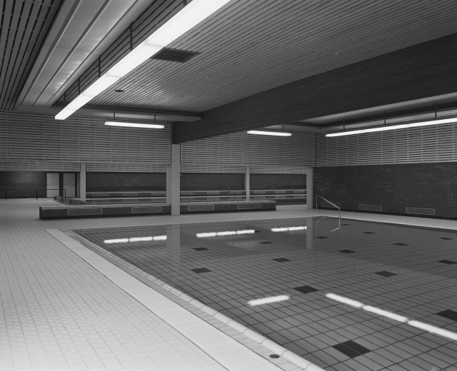 Indoor-Schwimmbad mit gefliestem Boden, Wänden und Deckenbeleuchtung, gefüllt mit klarem Wasser und umgeben von ein paar Gegenständen, die eine friedliche Atmosphäre schaffen.