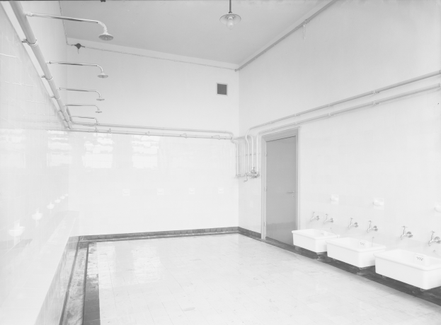 Ein Schwarz-Weiß-Foto einer öffentlichen Toilette mit Waschbecken, Wasserhähnen, Wänden, einer Tür, sichtbaren Rohren und einer Deckenlampe.